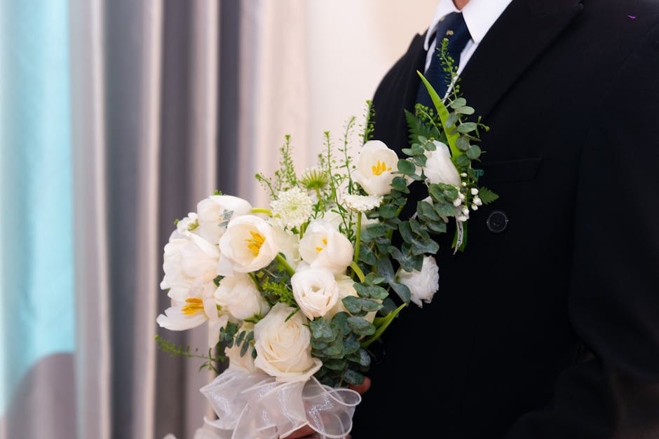 A person dressed in formal dark suit and white shirt holding a floral arrangement featuring white flowers, including roses and freesias, with green foliage accents. The bouquet is wrapped in white tul